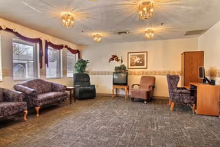 A cozy living room area with patterned purple upholstered chairs and a loveseat, a green recliner, a small wooden table with a TV, a framed picture on the wall, and a wooden desk with a computer. The room has three windows with purple valances and beige walls with a decorative border.