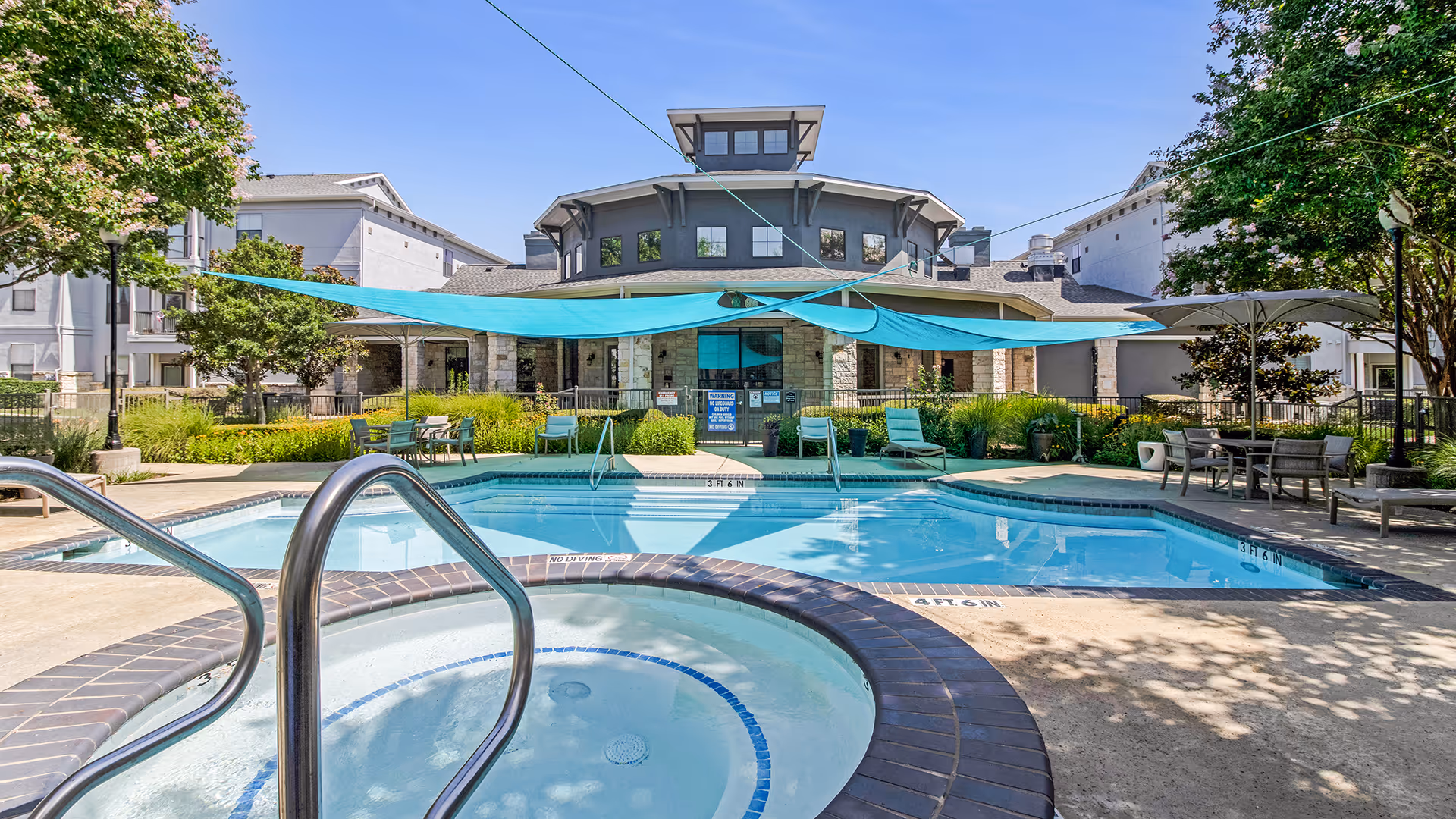Outdoor swimming pool and hot tub area at Chateau McKinney facility, surrounded by lounge chairs, tables, and shaded by blue fabric canopies with trees and a multi-story building in the background under a clear blue sky.