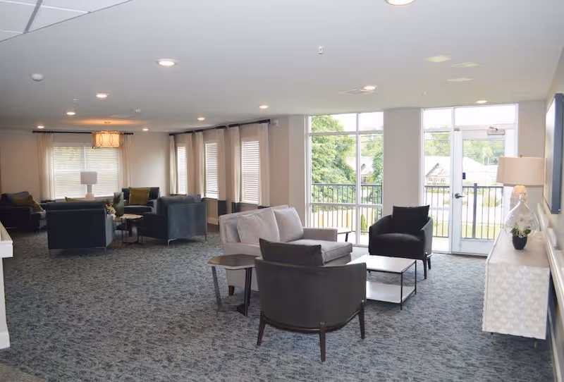 A spacious senior living facility common area with multiple seating arrangements including armchairs and sofas. Large windows and a glass door provide natural light and a view of greenery outside. The room is carpeted and decorated with lamps, curtains, and a sideboard with a plant and lamp on it.