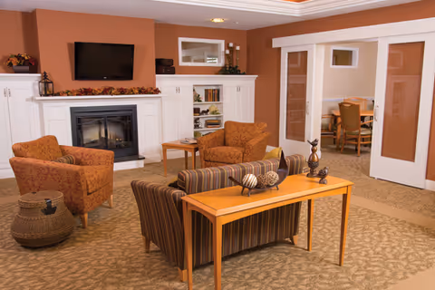 A cozy living room area with a striped sofa facing a white fireplace with a mounted flat-screen TV above it. Two patterned armchairs are arranged around a small wooden side table. A wooden console table behind the sofa holds decorative items. The room has warm earth tones with a carpeted floor and a doorway leading to a dining area with a table and chairs.