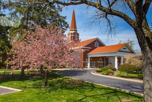 A red brick building with a tall steeple and a red roof, surrounded by green grass and trees, including a blooming pink cherry blossom tree in the foreground. The building has a covered entrance with white columns and a curved driveway leading up to it under a clear blue sky.