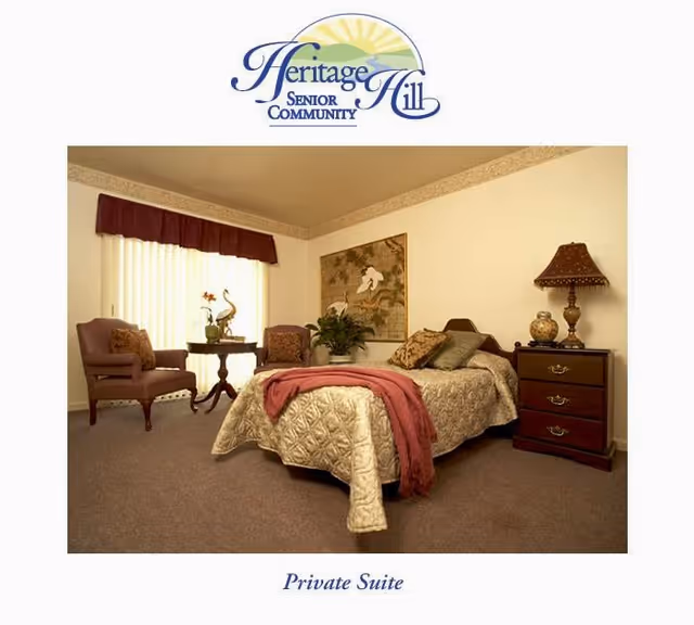 A cozy private suite in a senior living community featuring a single bed with a quilted bedspread and a red throw blanket, a wooden nightstand with a decorative lamp and vase, two upholstered chairs with a small round table between them, a large window with vertical blinds and a valance, and a framed artwork on the wall.