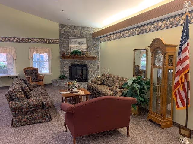 A cozy living room in a retirement community featuring floral patterned sofas, a maroon armchair, a wooden coffee table with a flower arrangement, a stone fireplace with a framed picture above it, an American flag, a tall wooden grandfather clock, a mirror, and two windows with light curtains.