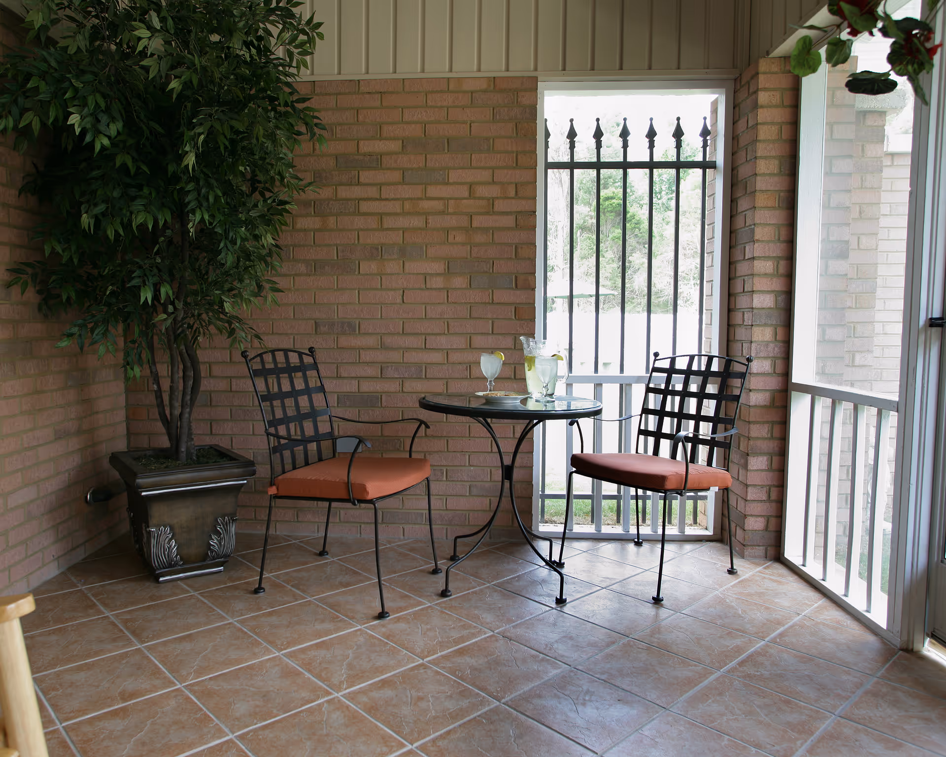 A small outdoor patio area with two metal chairs with orange cushions and a round glass-top table between them. On the table are two glasses of lemonade and a pitcher. There is a large potted plant to the left and a brick wall behind the seating area. The patio is enclosed with a metal gate and screened walls.