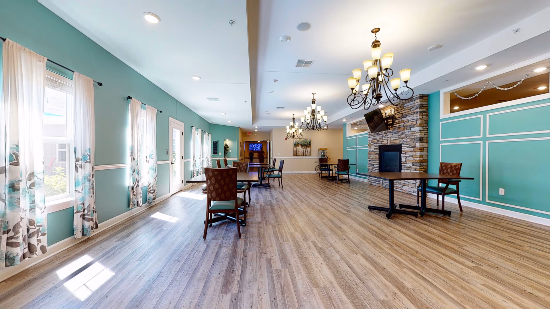 Spacious sunlit communal dining and lounge area with tables and chairs, chandeliers, and a stone fireplace.