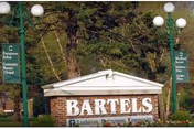 Outdoor view of a brick sign for Bartels Lutheran Retirement Community with green lamp posts and directional signs for Evergreen Arbor, Community Room, Chapel, Salon, Tables, Kitchen, and Linen Plan, surrounded by trees.