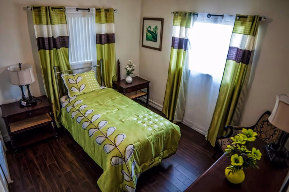 A small bedroom with a single bed covered in a green bedspread with a leaf pattern. The room has two windows with green, white, and brown striped curtains. There are two wooden nightstands on either side of the bed, each with a lamp. A framed picture hangs on the wall between the windows. A chair with a patterned cushion and a vase with yellow flowers on a dresser are also visible.