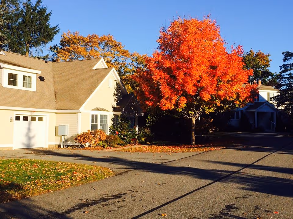 A residential building with beige siding and a light brown roof next to a tree with bright orange autumn leaves under a clear blue sky.