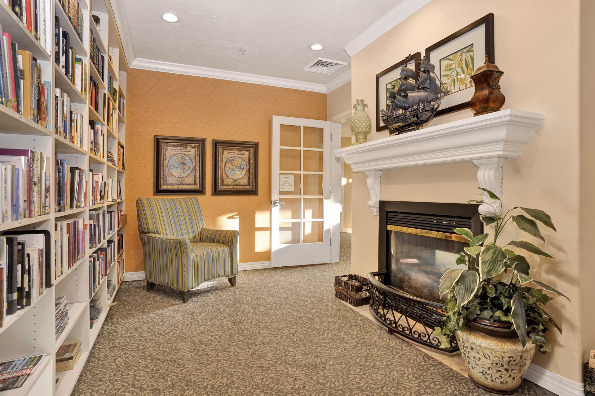 Cozy interior room with a striped armchair, a white bookshelf filled with books, a fireplace with decorative items on the mantel, two framed maps on an orange wall, and a potted plant beside the fireplace. A glass-paneled door is visible in the background.