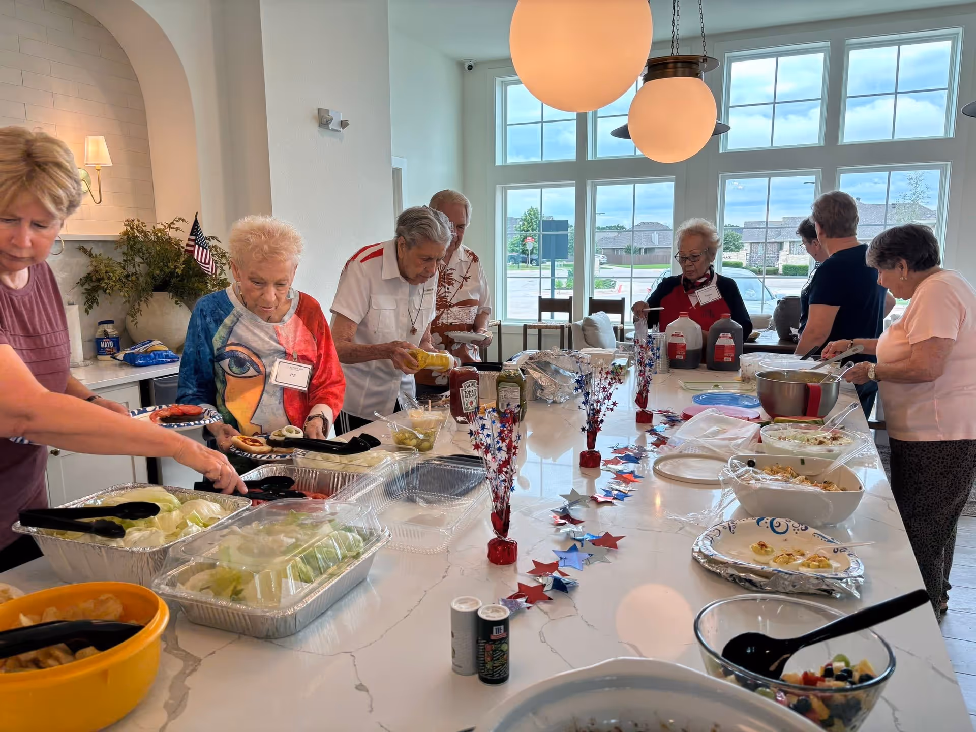 A group of elderly people serving themselves food from a buffet-style setup on a long kitchen island decorated with red, white, and blue star-themed decorations. The room is bright with large windows and hanging round light fixtures.