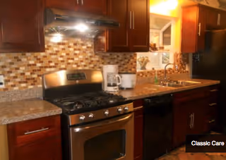 A kitchen with dark wooden cabinets, a stainless steel gas stove and oven, a coffee maker, a toaster, a double sink, and a mosaic tile backsplash in shades of brown and beige.