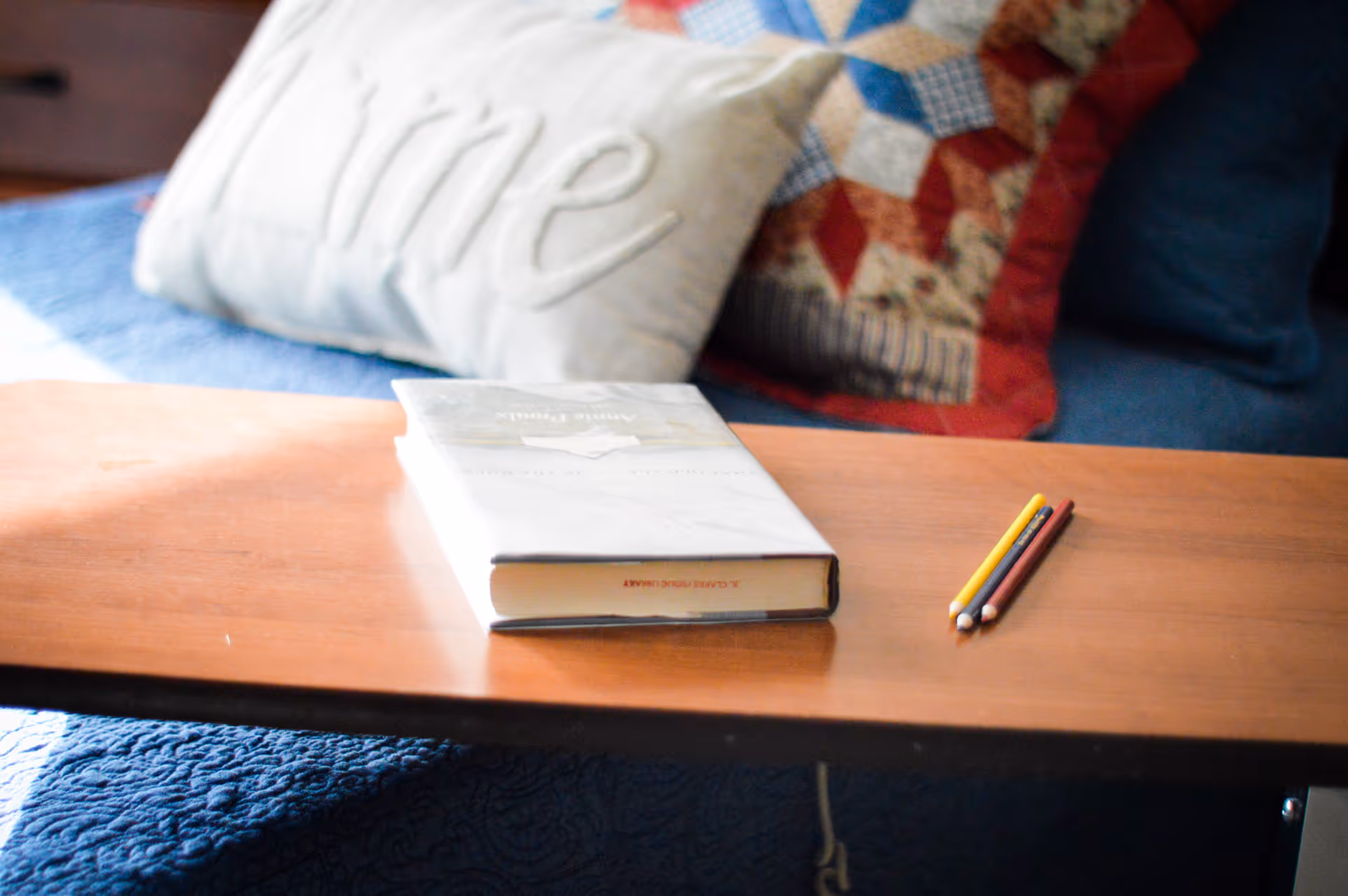 A wooden table with a closed book and three colored pencils on it. Behind the table is a blue quilted bedspread with a decorative pillow that has the word 'Home' embroidered on it and a colorful patchwork quilt.