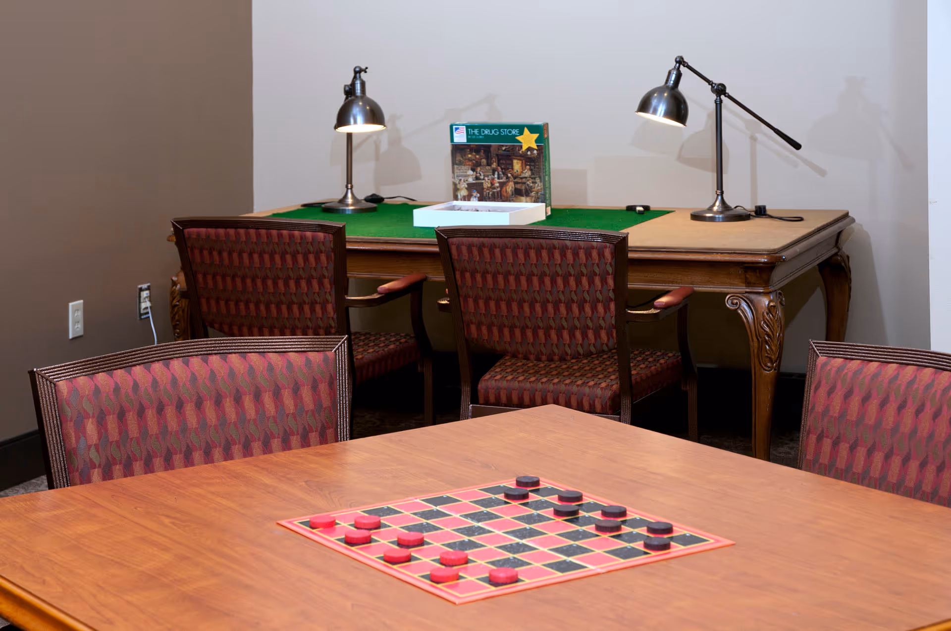 Activity room with a checkers board on a wooden table in the foreground and a green-covered game table with chairs and task lamps in the background.