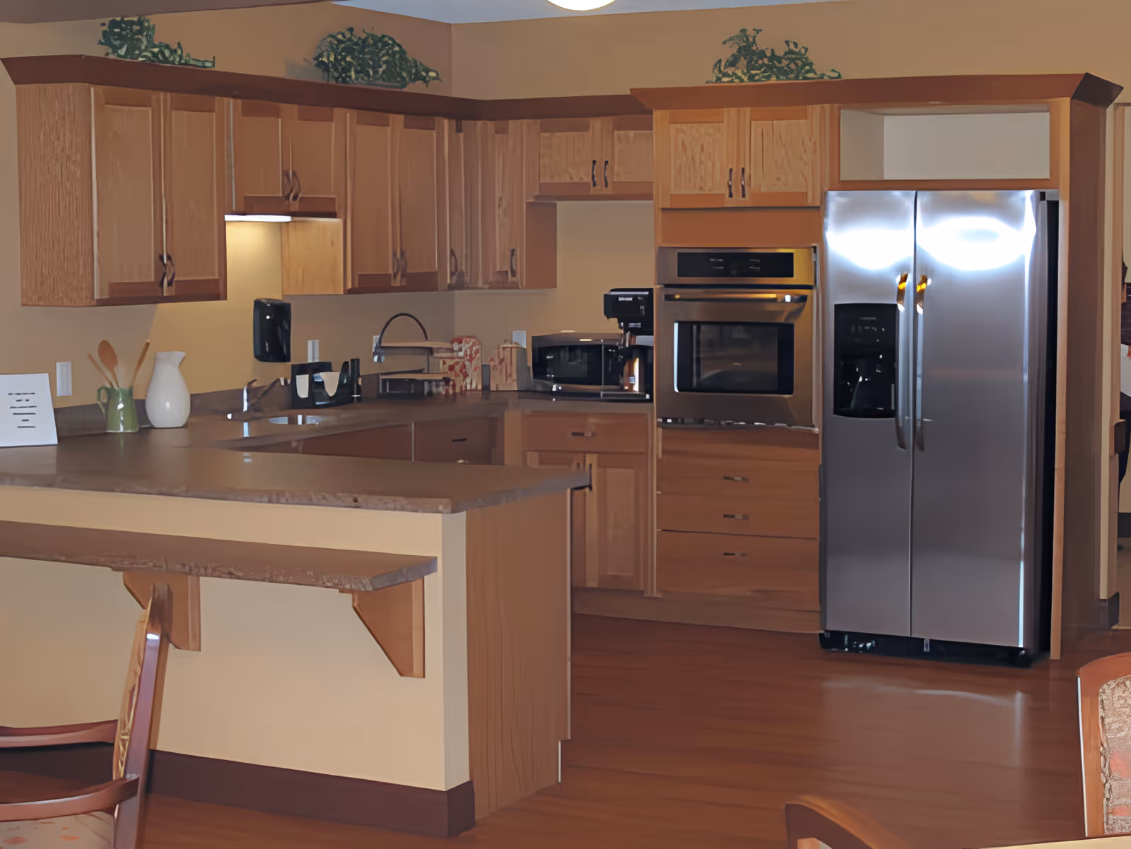 Open kitchen with wooden cabinets, stainless steel refrigerator and oven, and a countertop with seating.