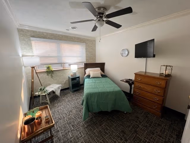 A small, neatly arranged bedroom with a single bed covered in a green blanket and a pillow. The room has a window with blinds, a ceiling fan, a wooden dresser with a TV mounted above it, a small side table with a lamp, and a floor lamp next to a small stool with a plant on it. The floor is carpeted with a patterned design.