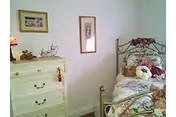 A cozy bedroom featuring a metal-framed bed with floral bedding and stuffed animals, a white dresser with decorative items on top, and two framed pictures hanging on the wall.