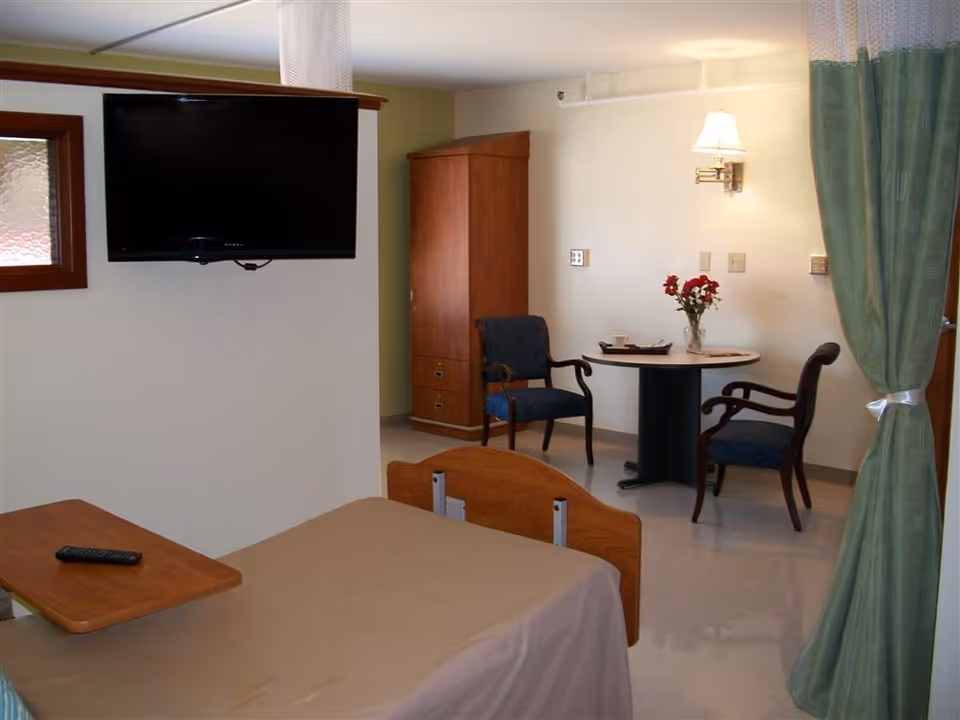 A senior living facility room with a hospital-style bed covered in a beige blanket, a wooden overbed table with a remote control, a wall-mounted flat-screen TV, a wooden cabinet, and a small round table with two upholstered chairs. A vase with red flowers is on the table, and a green curtain is tied back on the right side.