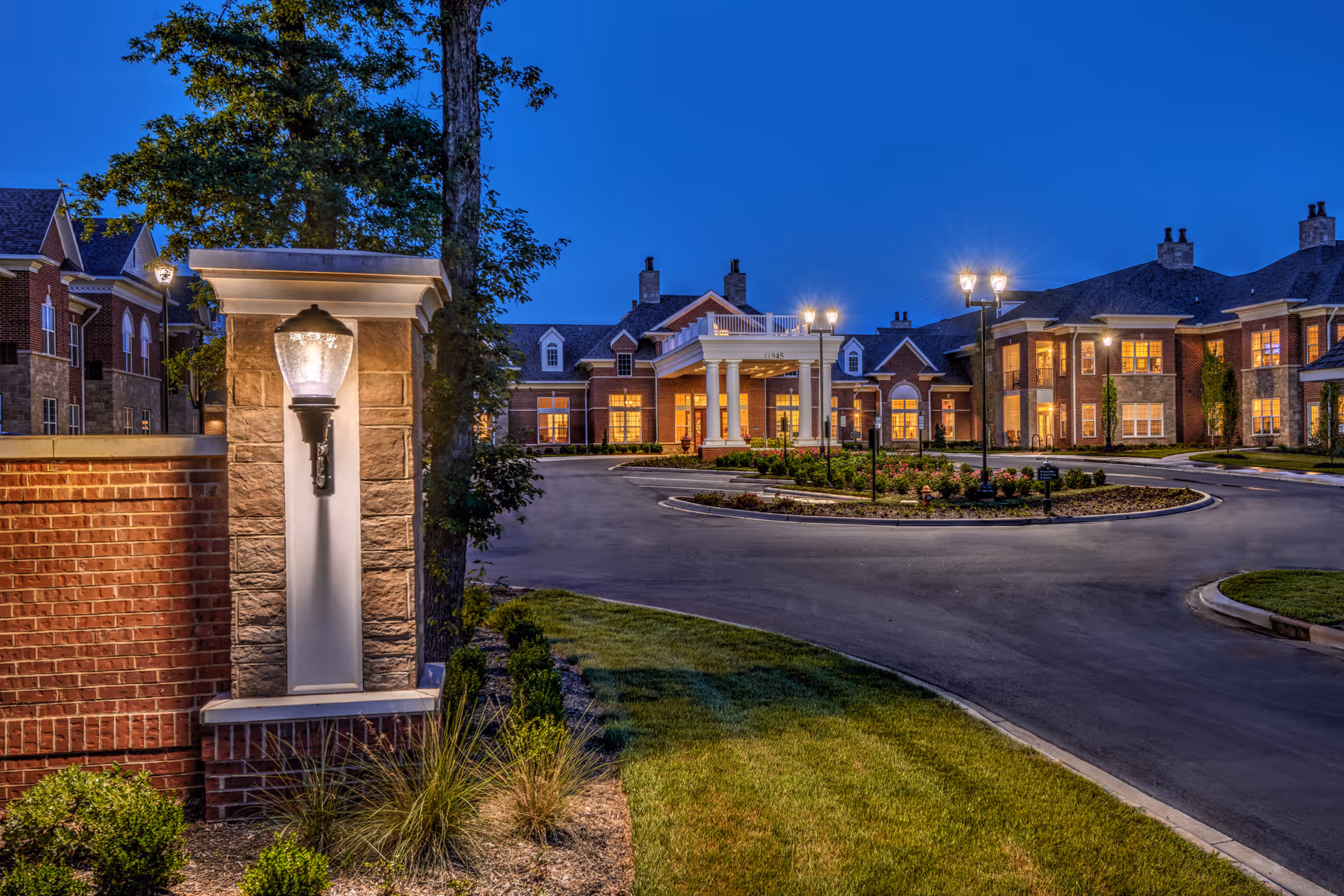 Evening view of the entrance to a senior living facility with a circular driveway, well-lit street lamps, and a large brick building with multiple windows and a covered entrance supported by white columns.