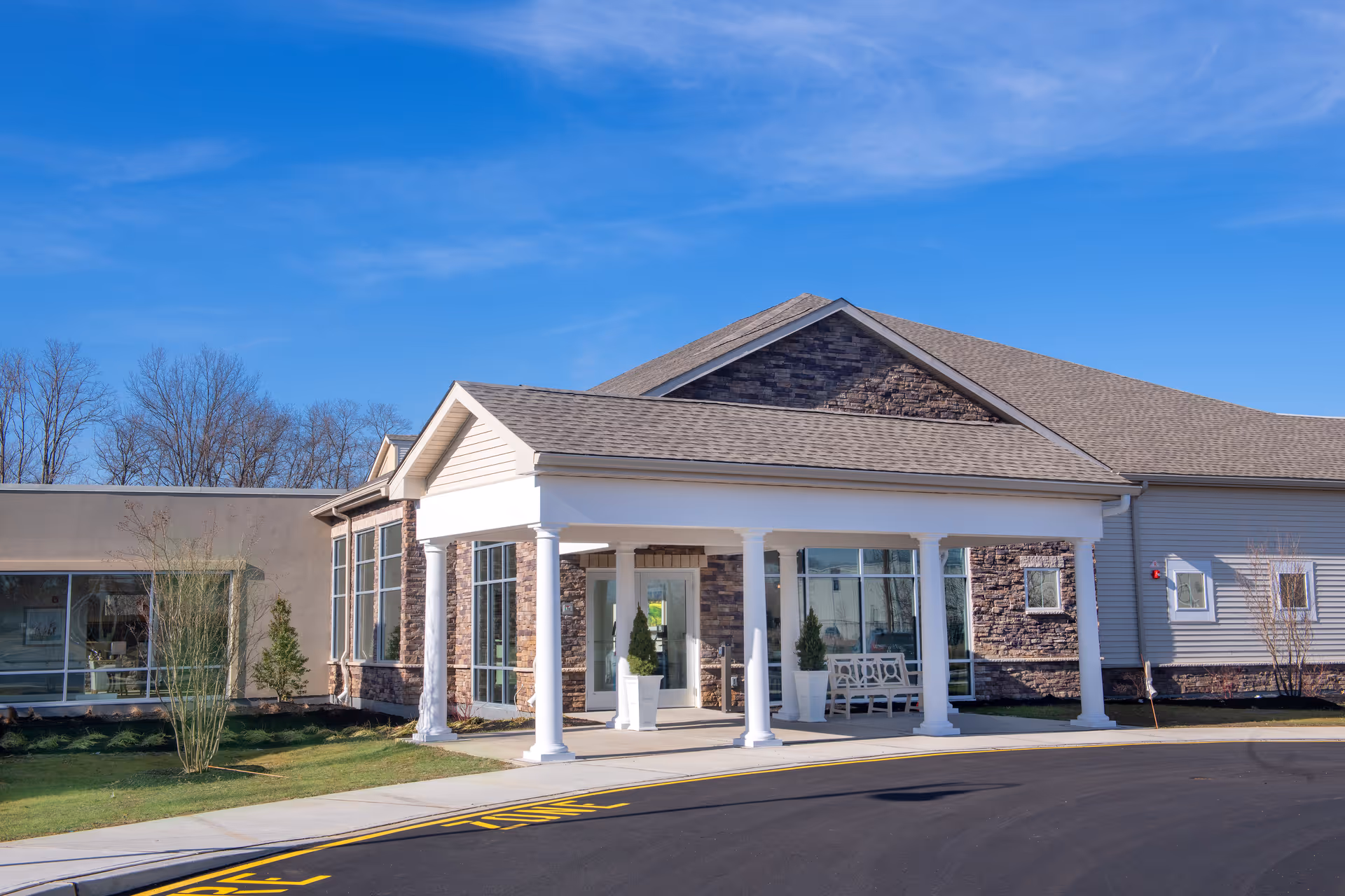 Exterior view of a senior living facility building with a covered entrance supported by white columns, large windows, and a bench near the entrance. The sky is clear and blue, and there are some small trees and shrubs around the building.