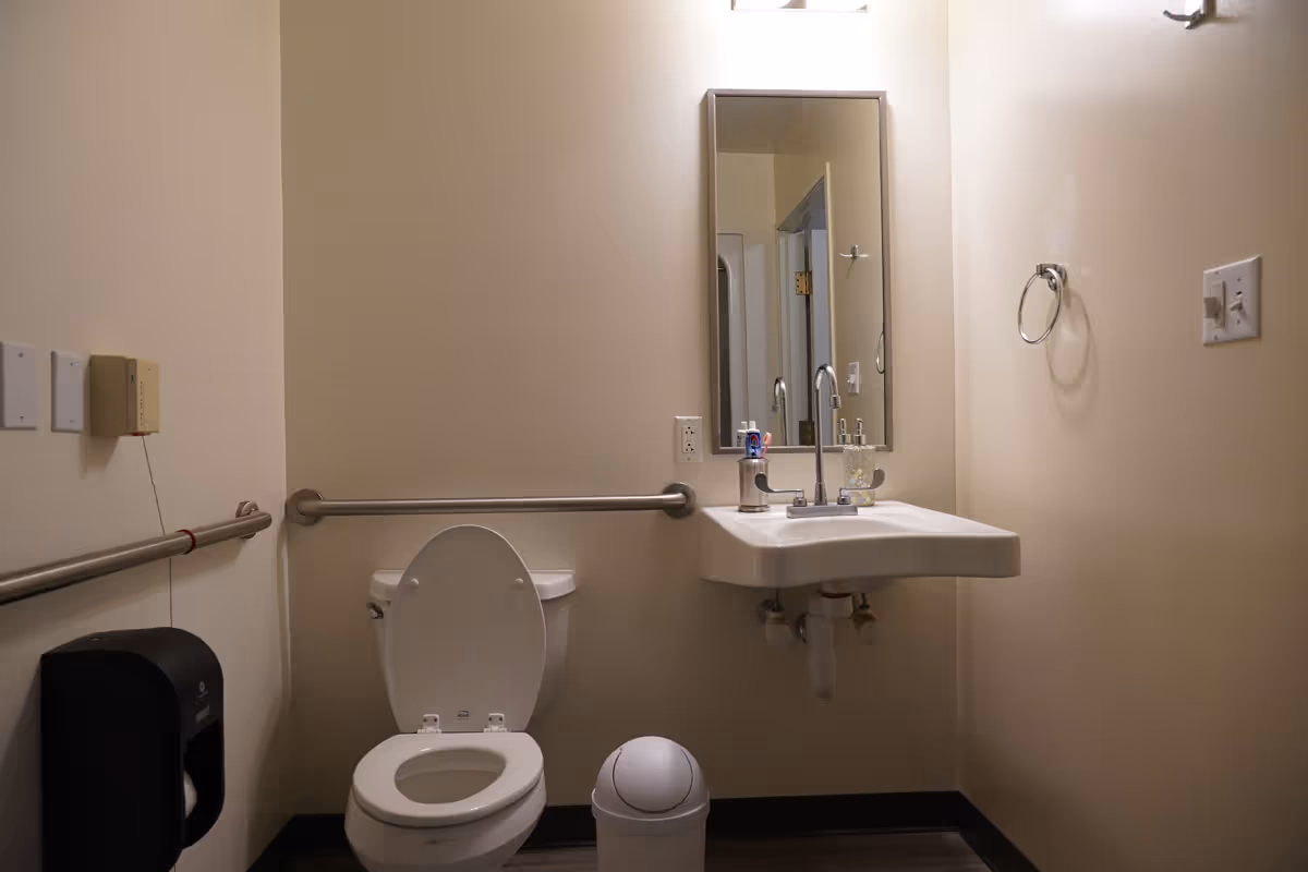A small, accessible bathroom with a toilet, a wall-mounted sink with a mirror above it, grab bars on the walls, a trash bin, and a toilet paper dispenser.