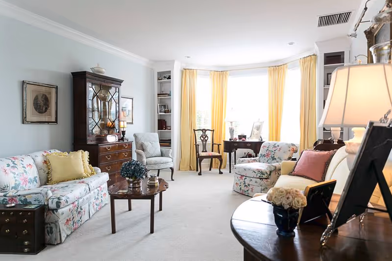 A bright and cozy living room with floral patterned sofas, a wooden coffee table, a vintage wooden cabinet, and a round side table with a lamp and flowers. The room has large windows with yellow curtains letting in natural light, and several chairs and framed pictures are visible.