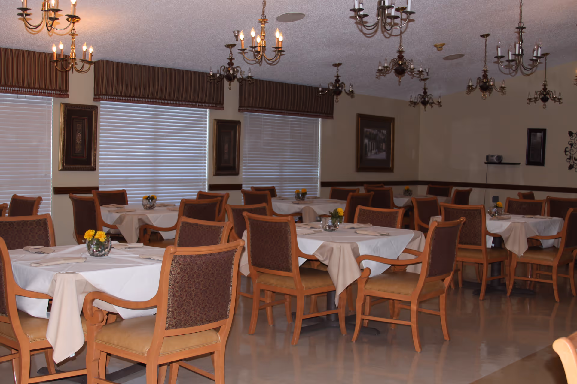 A dining room with multiple tables covered with white tablecloths and beige overlays. Each table has wooden chairs with cushioned seats and backs. Small flower arrangements are placed on each table. The room has several windows with blinds and valances, framed artwork on the walls, and multiple chandeliers hanging from the ceiling.
