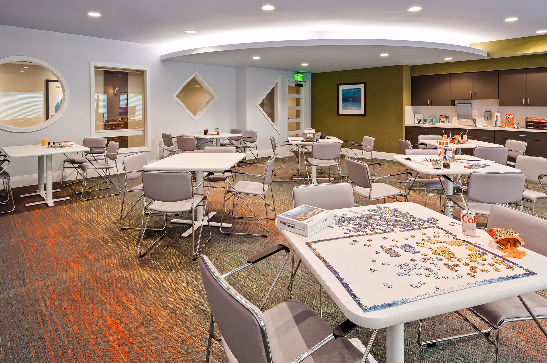 A spacious activity room with multiple tables and chairs arranged for group activities. One table in the foreground has a partially completed jigsaw puzzle, a Diet Coke can, and a bag of chips. The room has modern decor with a green accent wall, geometric windows, and a counter with cabinets along one side. The lighting is bright and the carpet has a colorful pattern.