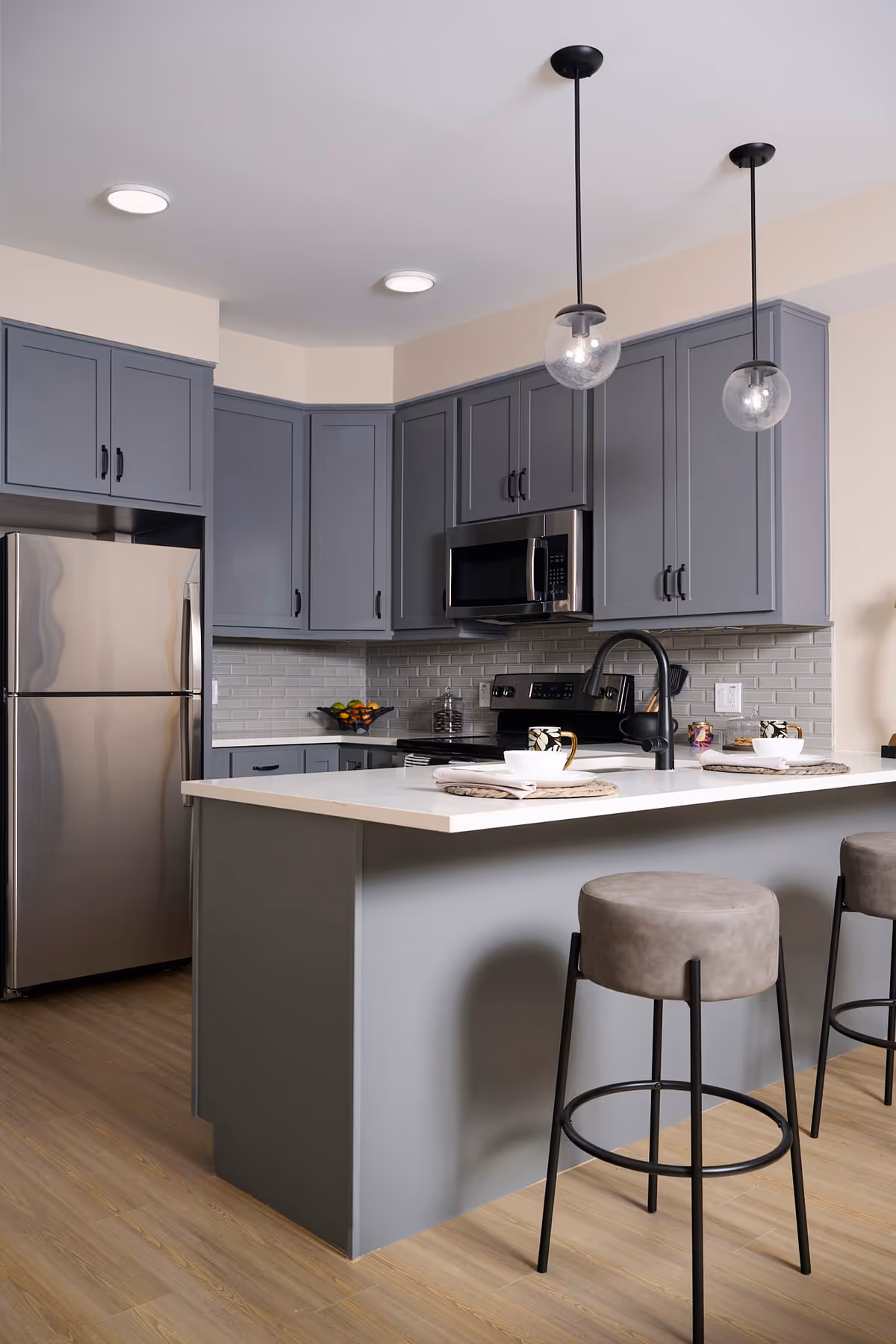 Modern kitchen with gray cabinets, stainless steel refrigerator and microwave, white countertop island with two place settings, and two gray bar stools. Two pendant lights hang from the ceiling above the island.