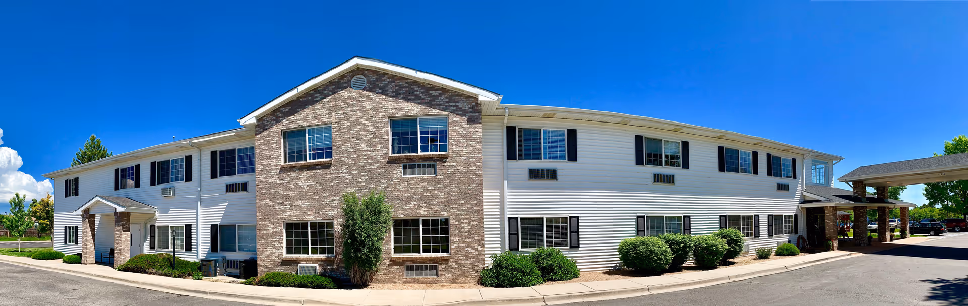 Exterior view of a two-story assisted living facility building with white siding and brick accents under a clear blue sky. The building has multiple windows with black shutters and a covered entrance on the right side. Shrubs and small trees are planted along the building's foundation, and a paved driveway runs in front.