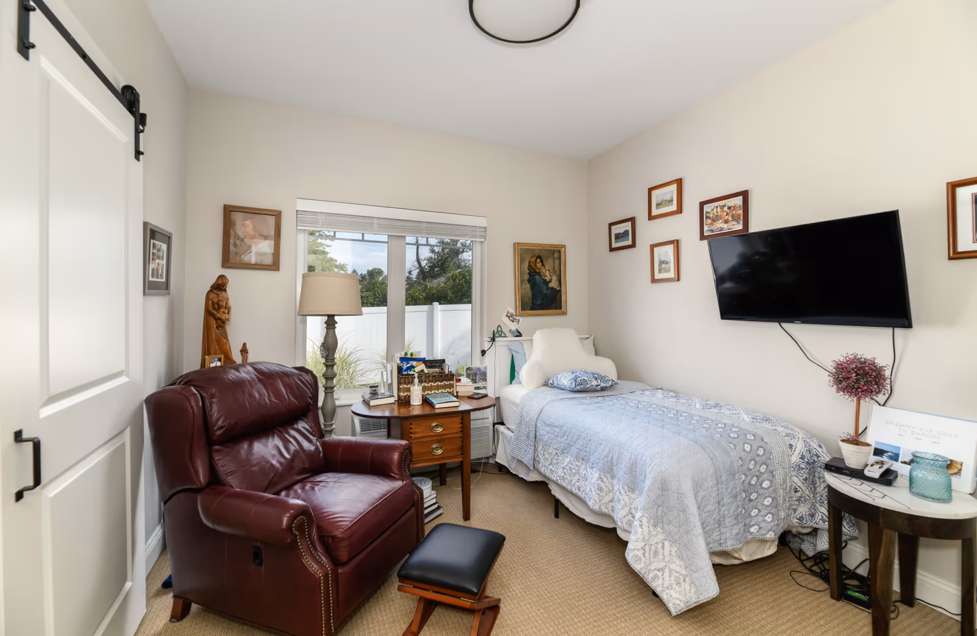 A cozy bedroom with a single bed covered in a blue and white patterned quilt. Above the bed is a wall-mounted flat-screen TV and several framed pictures. Next to the bed is a small round table with a potted plant, a candle, and some papers. A large burgundy leather armchair with a matching footrest is positioned near a window with white blinds. A wooden side table with a lamp and various items is placed between the armchair and the bed. The room has beige walls and carpeted flooring.