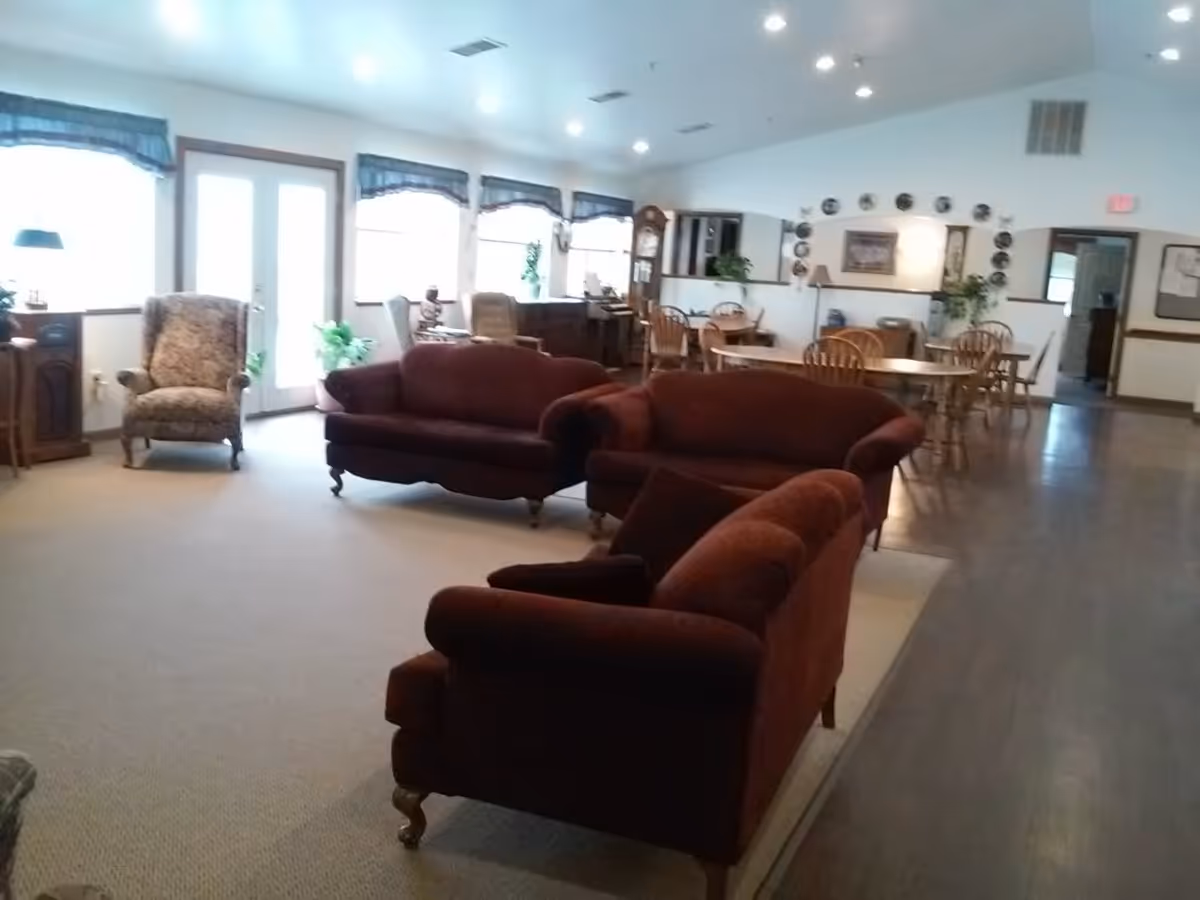 A spacious common area in a senior living facility with three maroon upholstered sofas arranged on a carpeted floor. In the background, there are wooden dining tables and chairs, a grandfather clock, and windows with blue valances letting in natural light. The room has a mix of carpet and wood flooring and is well-lit with ceiling lights.