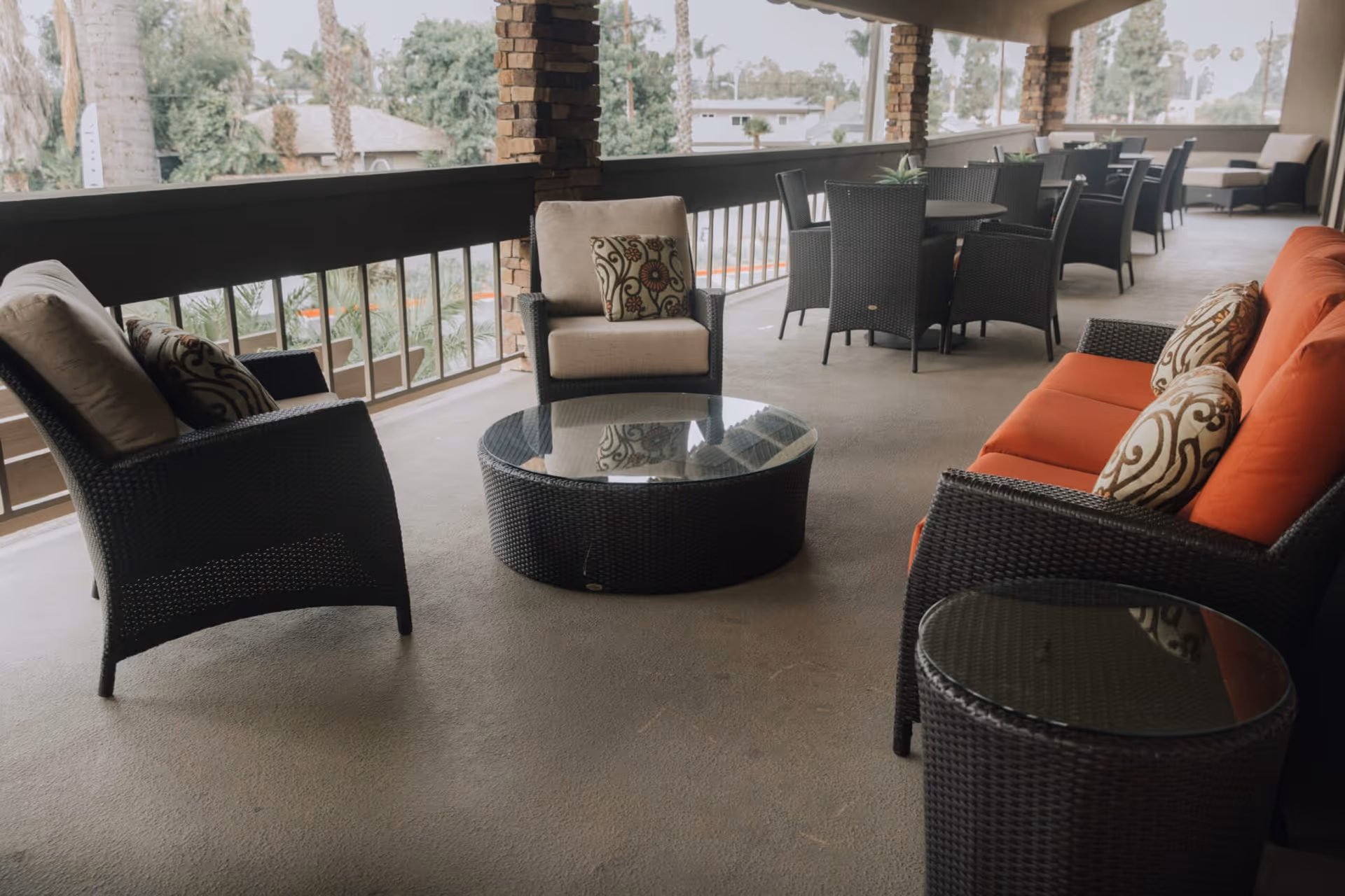 Outdoor covered patio area with wicker furniture including cushioned chairs, a sofa with orange cushions, and glass-topped tables. The patio overlooks trees and nearby houses.