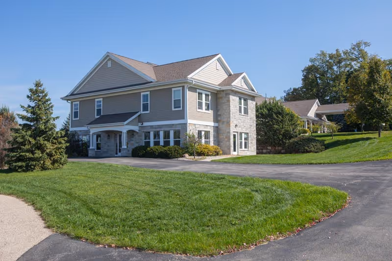 Two-story beige assisted living building with stone accents, surrounded by green lawn and a curved driveway under a clear blue sky.