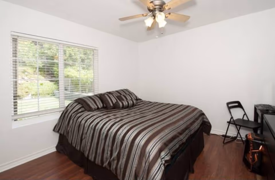 A small bedroom with a striped bedspread, window, ceiling fan, and folding chair.