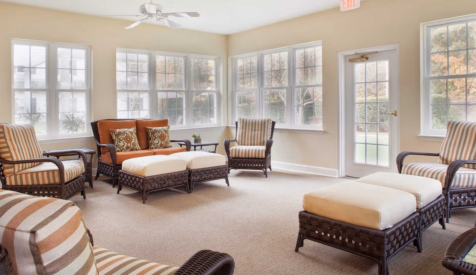 Bright communal sitting room with wicker-style chairs, cushioned ottomans, and large windows overlooking the grounds.