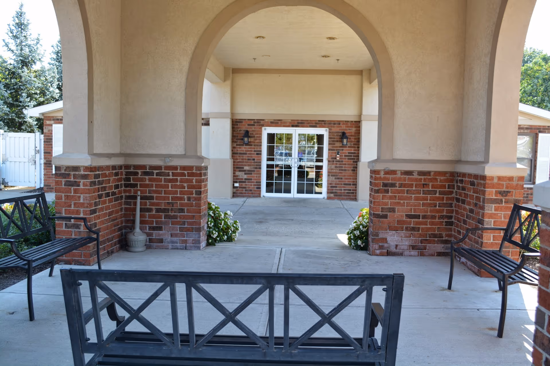 Covered outdoor seating area with black metal benches and brick pillars, leading to a building entrance with glass double doors and beige walls at Riverwalk Village.