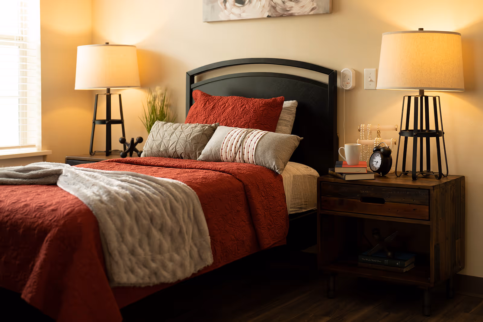 A cozy bedroom with a neatly made bed featuring a red quilt and multiple pillows. There are two bedside tables, each with a lamp. One table has a small plant and decorative items, while the other has books, a white mug, an alarm clock, and a beaded decoration. Soft natural light comes through a window with blinds.