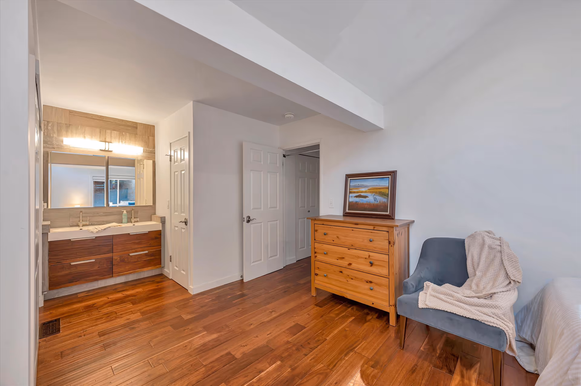 Interior view of a senior living facility room with wooden flooring, a wooden dresser with a framed painting on top, a blue upholstered chair draped with a beige blanket, and a bathroom vanity with two sinks and a large mirror. Two white doors are visible, one open and one closed.