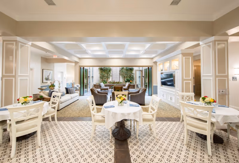 Bright communal dining and seating area with round tables and chairs in the foreground and sofas and a TV-lit seating area opening to a small patio garden.