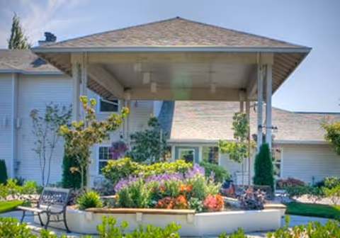 Covered porte-cochère entrance to a senior living building surrounded by landscaped flowerbeds and benches.