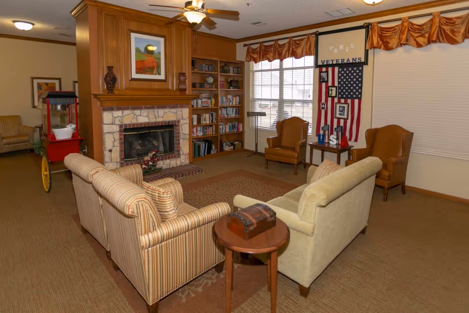 A cozy senior living facility common area with a stone fireplace surrounded by wooden paneling and built-in bookshelves. The room features two striped sofas facing the fireplace, a small round wooden table with a decorative chest, two brown leather armchairs near a window with blinds and valances, and a patriotic display with an American flag and the words 'Paris Veterans USA'. A popcorn machine is visible in the background.