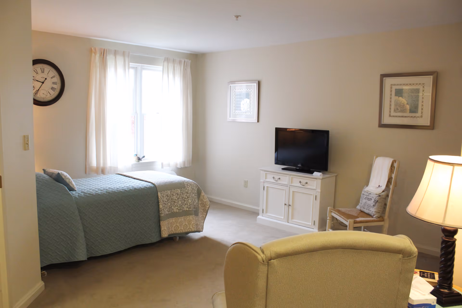 A cozy bedroom with a single bed covered in a blue quilt and a patterned blanket near a window with white curtains. A round wall clock is mounted on the left wall. Across from the bed is a white cabinet with a flat-screen TV on top. A wooden chair with a cushion and a folded blanket is next to the cabinet. A beige armchair and a table lamp with a twisted dark base are in the foreground. Two framed pictures hang on the wall above the TV and chair.