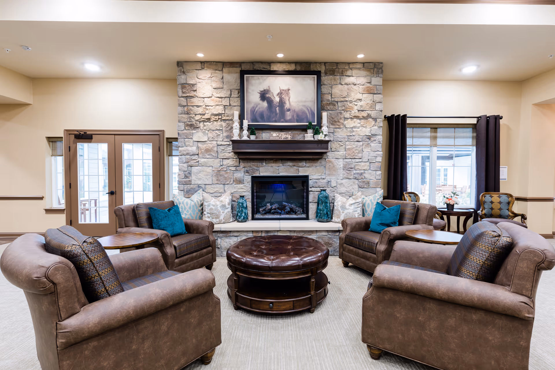 A cozy living room area with four brown leather armchairs arranged around a round, tufted leather ottoman. The room features a stone fireplace with a mantel decorated with candles, greenery, and a framed picture of two horses. There are windows with dark curtains on either side of the fireplace, and a door with glass panels on the left side of the room.