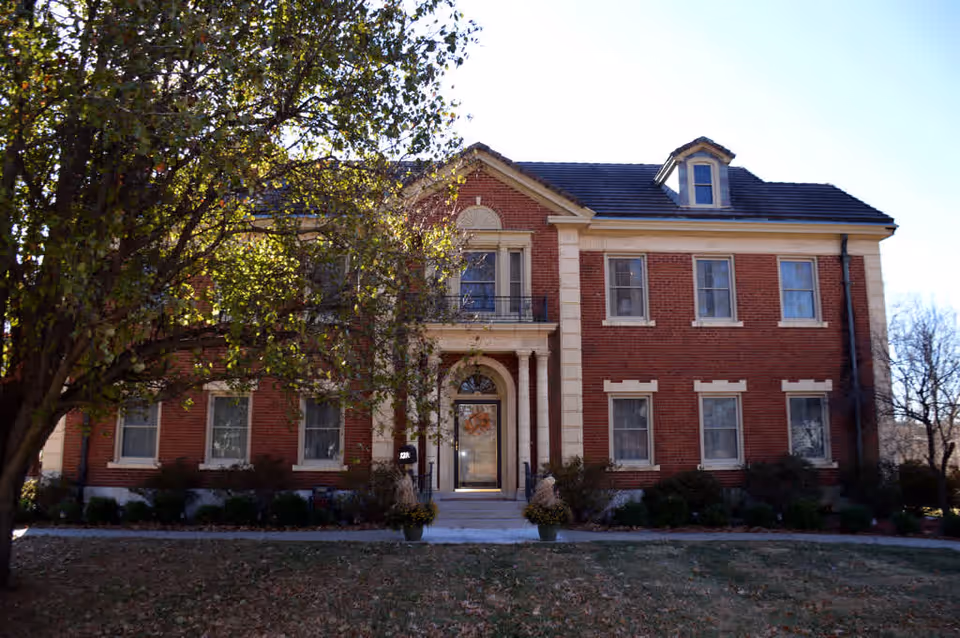 Two-story red brick building with a central columned entrance and dormer, seen from the front with a large tree on the left and a lawn in foreground.