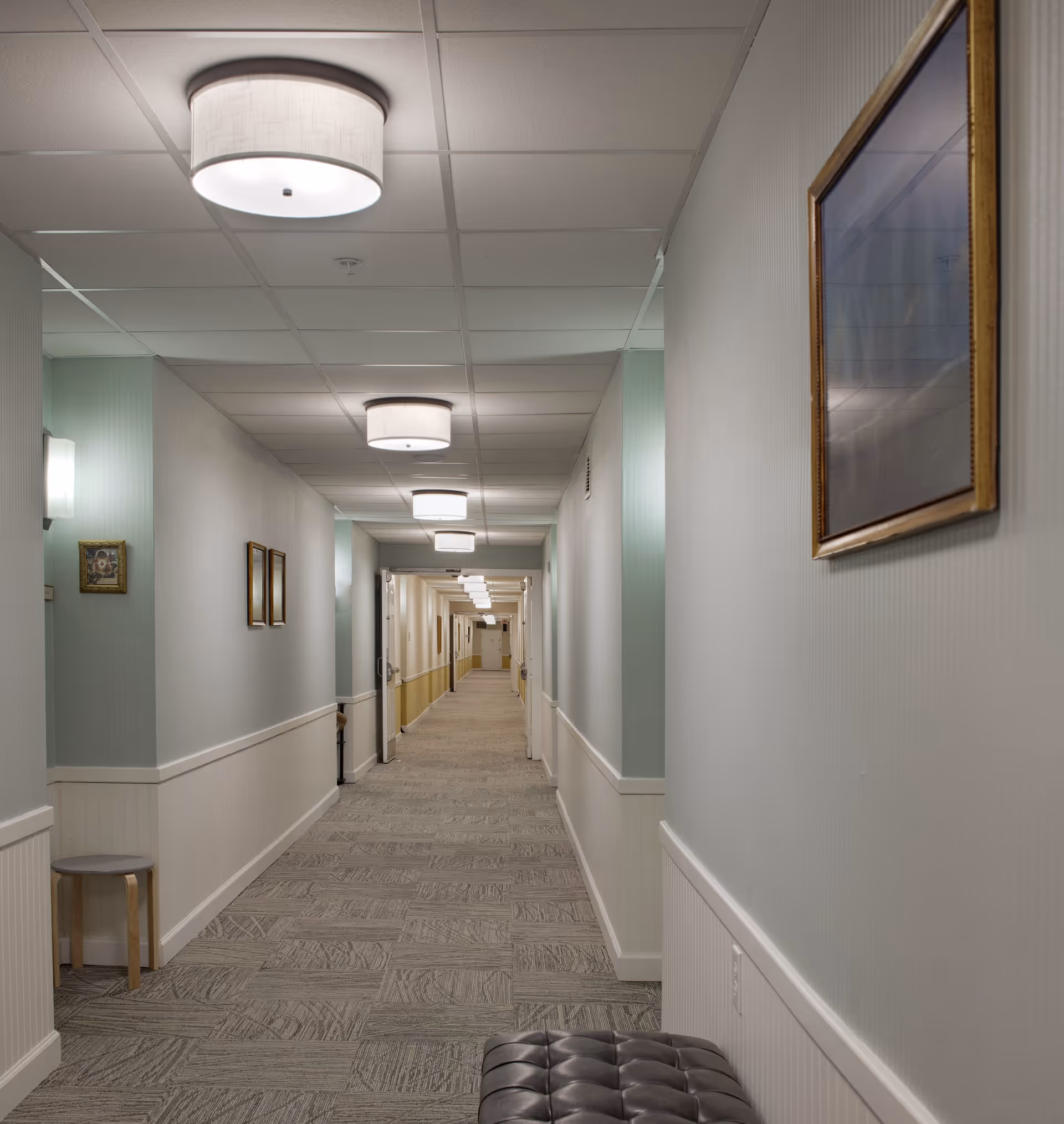 A long, well-lit hallway in a senior living facility with light-colored walls, carpeted floor, and ceiling lights. There are framed pictures on the walls and a small bench on the right side near the foreground.