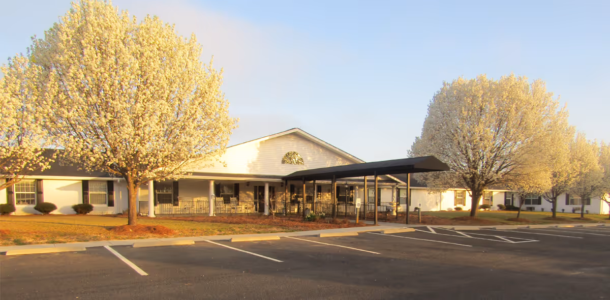 Front exterior of a single-story white senior living building with a covered entrance, blossoming trees, and empty parking spaces.