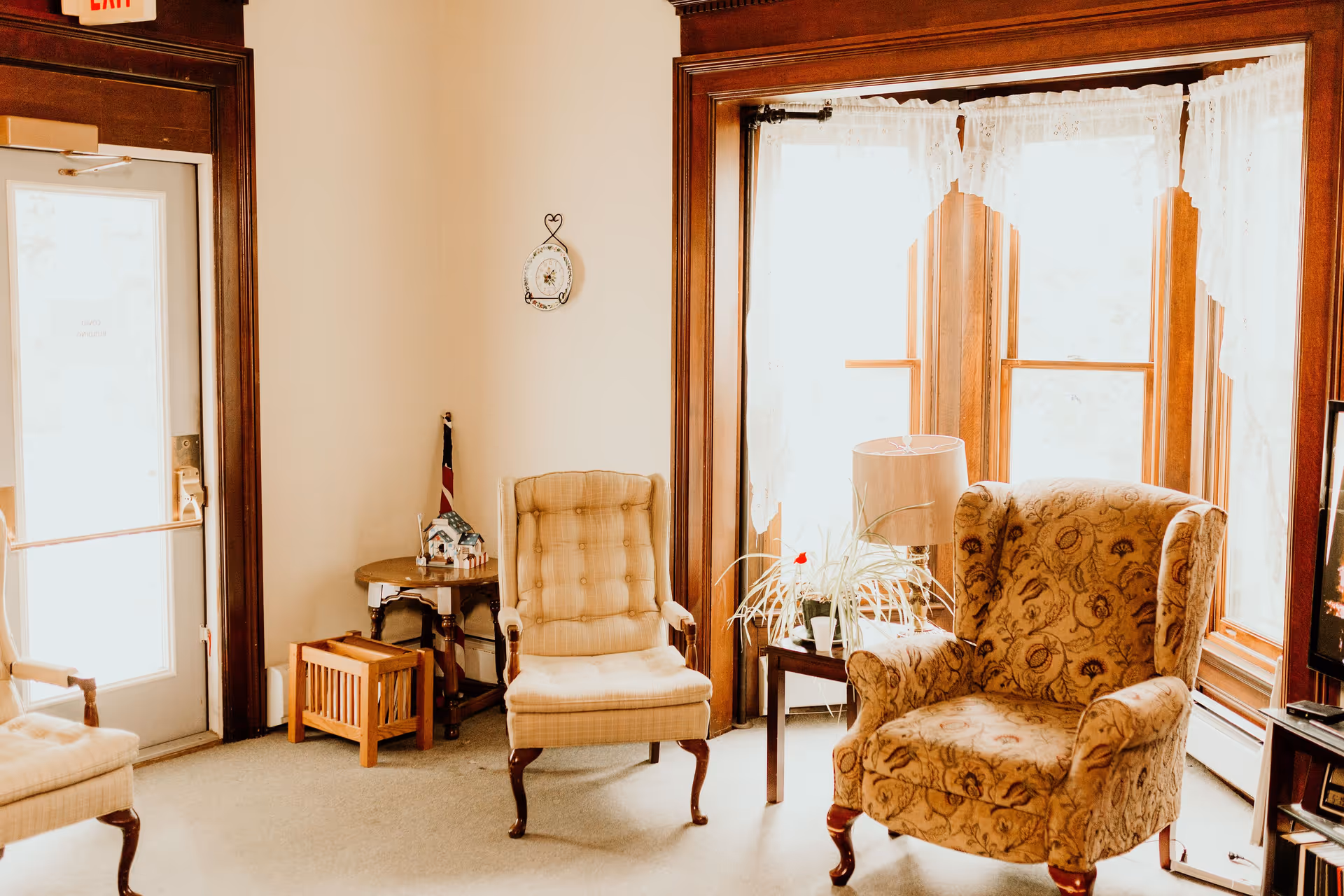 A cozy living room area with two upholstered armchairs, a small wooden side table with a lamp and a potted plant, large windows with lace curtains letting in bright sunlight, and a door with an exit sign above it.