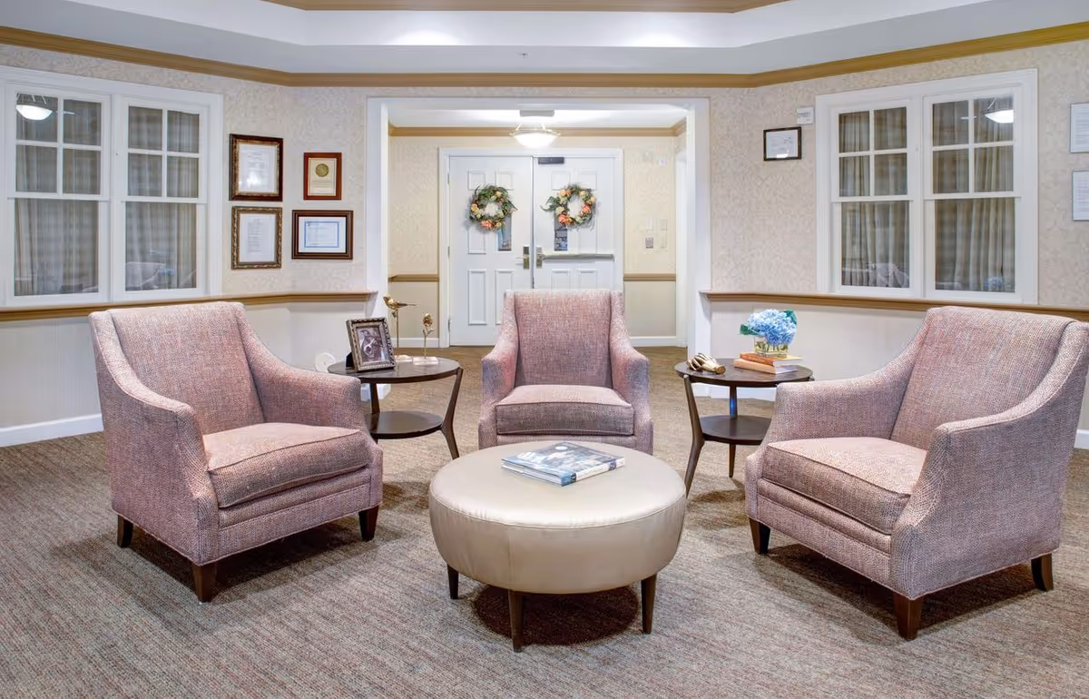 A cozy sitting area in a senior living facility with three upholstered armchairs arranged around a round beige ottoman. Two small side tables hold decorative items including a framed photo, a small sculpture, books, and a vase with blue flowers. The background features double doors adorned with floral wreaths and framed certificates on the walls.