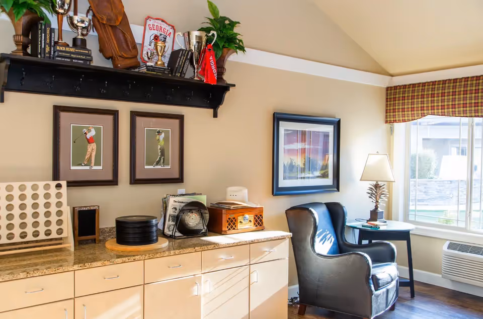 A cozy interior corner of a senior living facility with a black leather armchair next to a small round table holding a lamp. Above a beige countertop with cabinets, there are two framed golf-themed pictures on the wall and a black shelf displaying trophies, books, and decorative items. A large window with a plaid valance lets in natural light.