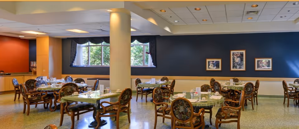 Dining room with multiple round tables set for meals, floral-upholstered chairs, large windows, and framed artwork on the wall.